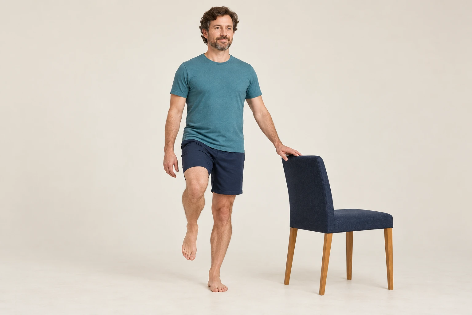 A man balances on one flat foot with the other foot lifted and fingertips on a chair beside him